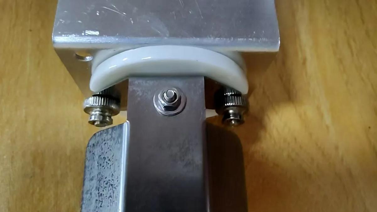 Close-up of the base joint on a Shield unit: the reflector shield&rsquo;s end tab clamped between the white lampholder and the magnetic bracket plate by two knurled thumb nuts, with the bracket&rsquo;s own centre bolt between them