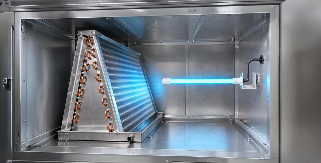 Cutaway view inside a stainless air handler: the finned evaporator coil stands on the left and a BioShield UV-C lamp is bracketed to the duct wall on the right, its blue light washing across the coil face.