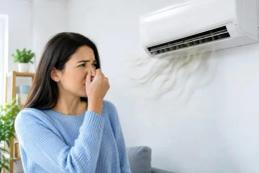 A woman pinching her nose at the stale air coming from a wall-mounted air conditioner.